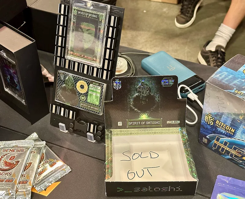 A booth table displaying framed Spirit of Satoshi cards beside a handwritten “Sold Out” sign