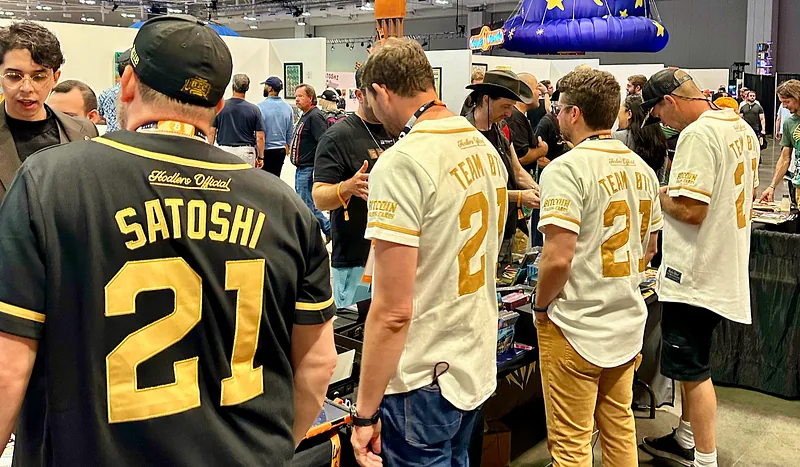 Collectors in Bitcoin Trading Cards jerseys gathered around the booth, one of them wearing a Satoshi 21 jersey
