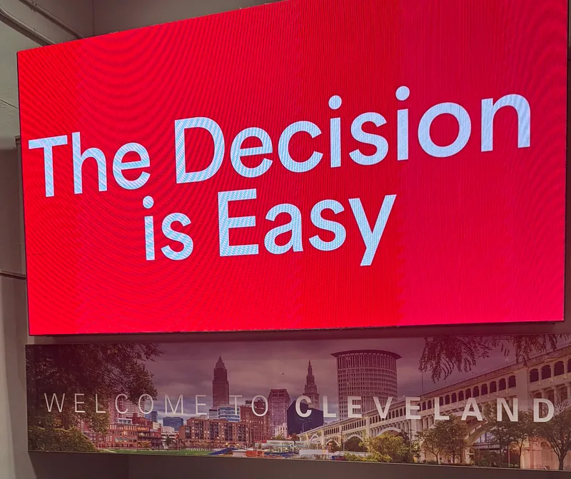 A large red PSA advertising screen in an airport reading “The Decision is Easy”, above a “Welcome to Cleveland” panel