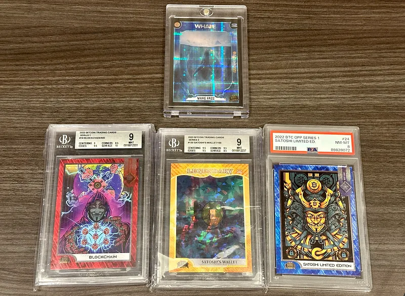 Three slabbed Bitcoin Trading Cards on a table, two of them graded 9 by Beckett and one a Series 1 Satoshi Limited Edition