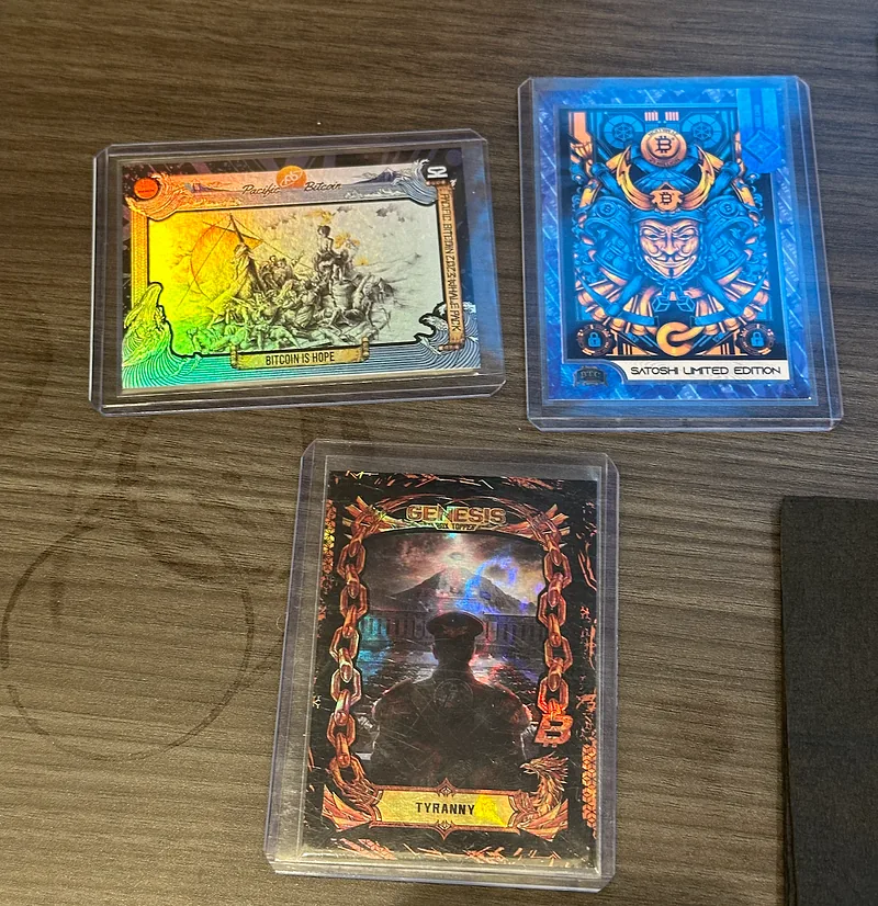Three Bitcoin Trading Cards on a wooden table: a Genesis Tyranny card below, and a Satoshi Limited Edition and a second card above it