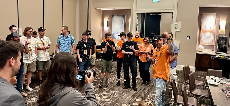 A crowd standing in a hotel room listening to Alladan, in an orange shirt, give a speech